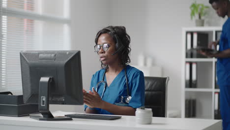 African-Female-medical-assistant-wears-white-coat-headset-video-calling-distant-patient-on-computer.-Doctor-talking-to-client-using-virtual-chat-computer-app.-Telemedicine-remote-healthcare